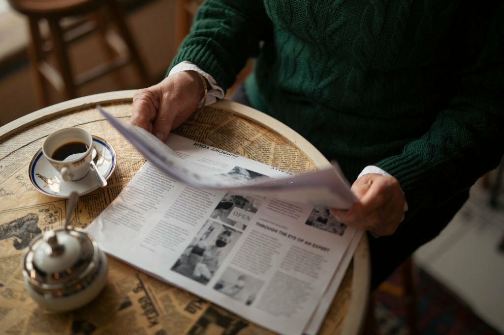 Midsection of older woman with newspaper reading theater reviews and sitting by table in coffee shop and having morning coffee, cut out.