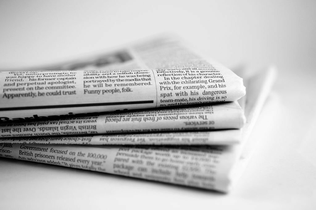 A close up black and white image of a stack four broadsheet newspapers with English text, casually piled up on a plain white background. The macro image is shot with a shallow depth of field, with only generic text visible. With copyspace.