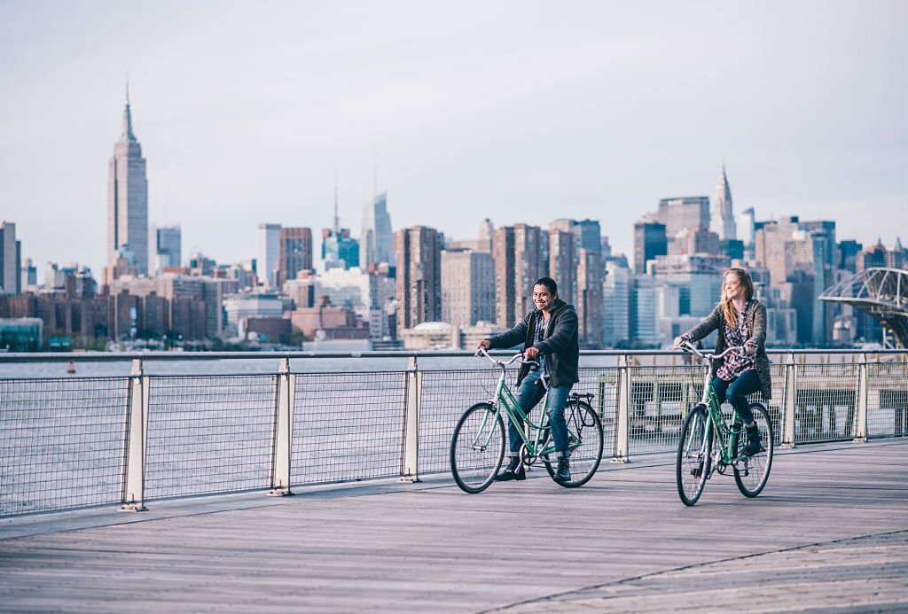 Young couple is cycling on a pier. They are looking around. New York in the back.