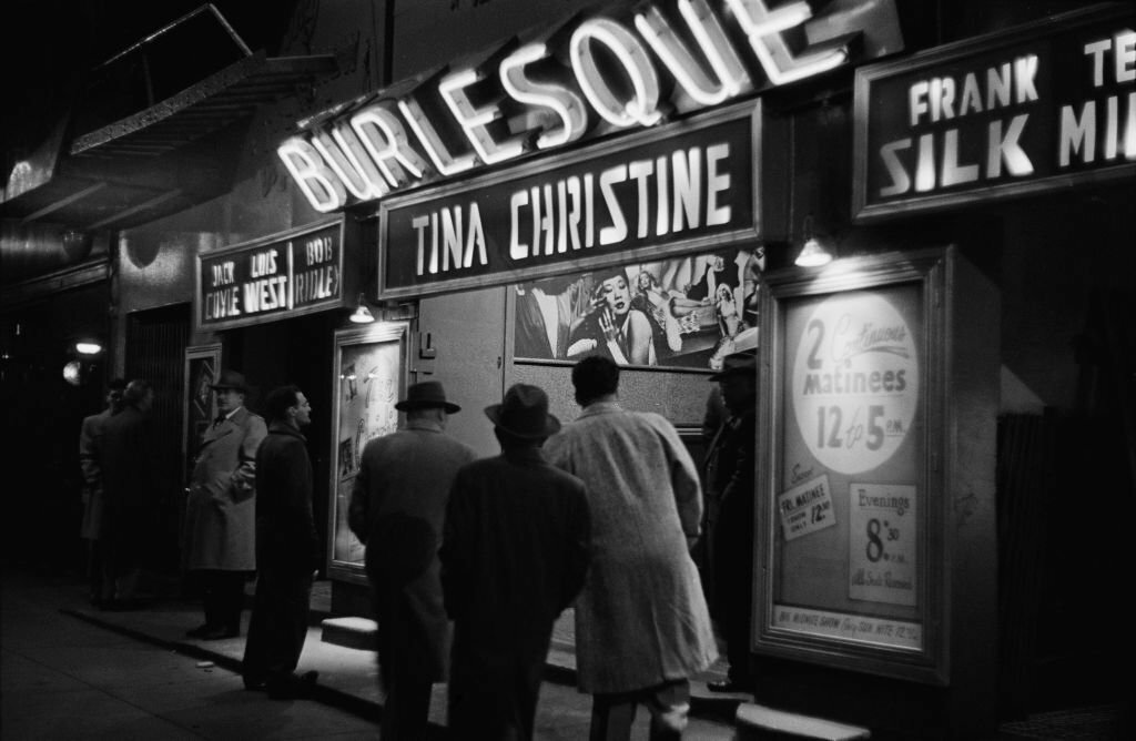 men outside a burlesque club, with the names of 'Jack Coyle', 'Luis West', 'Bob Ridley', 'Tina Christine', and 'Frank Silk' illuminated below an illuminated 'burlesque' sign, United States, 1953. A framed notice advertises '2 continuous matinees, 12 to 5 pm'. 
